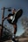 A person in athletic attire is captured from a low angle while kicking towards the camera, creating a dynamic perspective. The person is wearing sunglasses and standing on a bridge with rope railings, surrounded by trees with clear skies above.