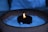 A candle with a steady flame is set inside a circular holder, surrounded by small white pebbles or salt within a rustic ceramic pot. The scene is softly illuminated with the surrounding area appearing dimly lit.