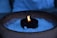 A candle with a steady flame is set inside a circular holder, surrounded by small white pebbles or salt within a rustic ceramic pot. The scene is softly illuminated with the surrounding area appearing dimly lit.