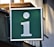 A large green signboard with a white lowercase 'i' symbol, often representing information. It is mounted on the side of a building, with a small plant visible in the upper corner.