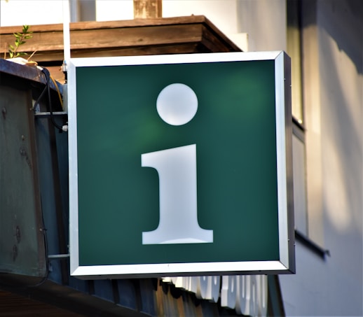 A large green signboard with a white lowercase 'i' symbol, often representing information. It is mounted on the side of a building, with a small plant visible in the upper corner.