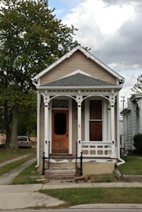 A small, vintage-style house with a wooden front door and a narrow porch supported by decorative trim. The siding of the house is light brown with white accents. A large tree stands to the left, and a driveway with a parked car extends to the side of the house. The sky is partly cloudy, creating a tranquil atmosphere.