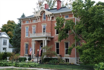A large, elegant brick house features classic architectural elements, including tall windows, decorative lintels, and a symmetrical facade. The house is adorned with two flags on the porch, surrounded by a well-maintained garden and mature trees. A wrought iron fence borders the front yard, enhancing the traditional aesthetic of the property.