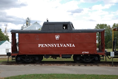 A vintage red caboose with 'PENNSYLVANIA' written on its side is set on railway tracks. The background includes various industrial structures and blue sky with clouds.