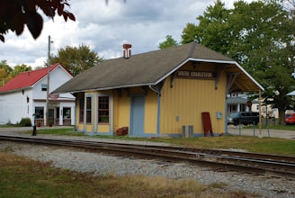 A yellow wooden train station building marked 'South Charleston' stands beside a railway track, with a historical and small-town atmosphere. The structure has a pitched roof and is accompanied by a stop sign and a few surrounding trees and buildings. In the background, there is another white building with a red roof, indicative of a quaint, rural setting.