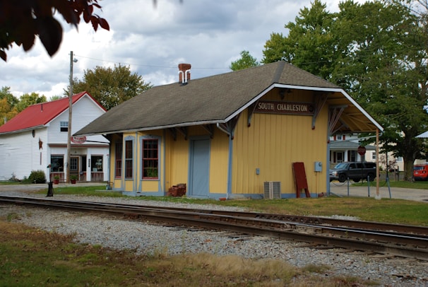 A yellow wooden train station building marked 'South Charleston' stands beside a railway track, with a historical and small-town atmosphere. The structure has a pitched roof and is accompanied by a stop sign and a few surrounding trees and buildings. In the background, there is another white building with a red roof, indicative of a quaint, rural setting.