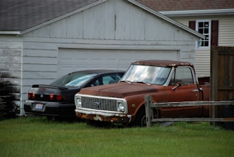 An old, rusted pickup truck is parked alongside a black car in front of a white garage. The truck is brown with noticeable rust patches, and the grass around it is slightly overgrown. A wooden fence partially encloses the area, and a house with brown shutters can be seen in the background.