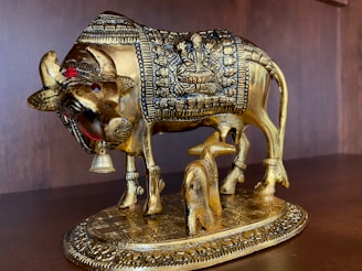 A golden sculpture of a cow with intricate carvings on its body, depicting a deity figure. It stands on a decorated base, with smaller details like a bell around its neck. The statue is positioned on a wooden surface or shelf.