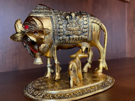 A golden sculpture of a cow with intricate carvings on its body, depicting a deity figure. It stands on a decorated base, with smaller details like a bell around its neck. The statue is positioned on a wooden surface or shelf.