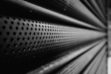A close-up perspective of a perforated metal surface with numerous circular holes, creating a repetitive pattern. The texture and depth of field emphasize the industrial nature of the material, with a play of light and shadow across the surface.