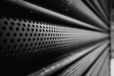 A close-up perspective of a perforated metal surface with numerous circular holes, creating a repetitive pattern. The texture and depth of field emphasize the industrial nature of the material, with a play of light and shadow across the surface.