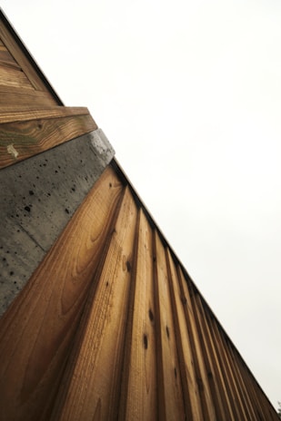 A diagonal view of a wooden and concrete structure, focusing on the texture and lines of the materials. The wooden panels are arranged vertically, showcasing a natural pattern and grain, while the concrete has a rough, pitted surface with some weathered areas.