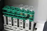 A set of four clear glass vials are placed in a green plastic rack. The rack is mounted on a larger beige plastic structure with a barcode label. Some of the vials contain a light-colored liquid.