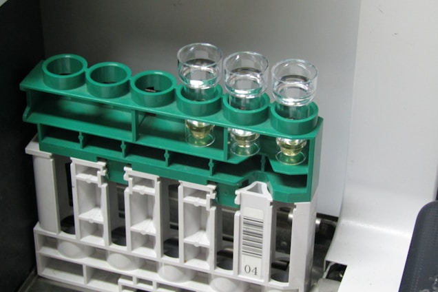A set of four clear glass vials are placed in a green plastic rack. The rack is mounted on a larger beige plastic structure with a barcode label. Some of the vials contain a light-colored liquid.