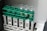 A set of four clear glass vials are placed in a green plastic rack. The rack is mounted on a larger beige plastic structure with a barcode label. Some of the vials contain a light-colored liquid.