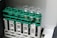 A set of four clear glass vials are placed in a green plastic rack. The rack is mounted on a larger beige plastic structure with a barcode label. Some of the vials contain a light-colored liquid.