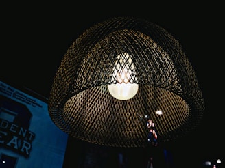 A warmly lit bulb is encased in a dark, mesh wire lampshade creating a cozy ambiance. The background is dimly lit with some text and a logo barely visible, adding an industrial and rustic aesthetic.