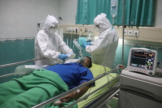 Medical professionals in full protective gear attend to a patient lying on a hospital bed. The patient is connected to medical equipment, including a heartbeat and oxygen monitor, while the healthcare workers appear to be providing care or administering a treatment.