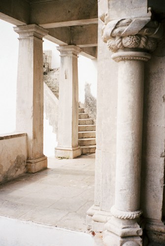 A stone staircase is partially visible beyond a series of Romanesque columns and arches. The architecture features detailed carvings and weathered surfaces, evoking a sense of antiquity. The light is soft and natural, creating gentle shadows on the stone floor.
