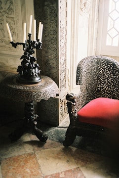 A richly detailed room featuring ornate furniture. A dark, intricately carved wooden table supports a tall, decorative candelabra with unlit candles. Next to it, a chair with elaborate patterns and a vibrant red cushion is set against an intricately carved wall panel.