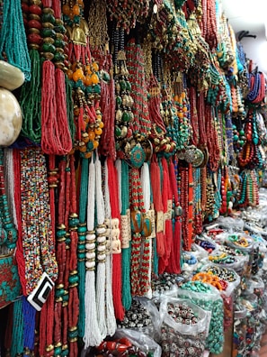 A vibrant display of colorful necklaces and beads, with shades of red, green, and gold dominating the intricate patterns. The necklaces are densely arranged, creating a rich tapestry of textures and materials. Located in a market setting, the variety and abundance of jewelry convey a sense of cultural richness.