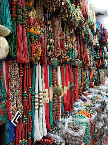 A vibrant display of colorful necklaces and beads, with shades of red, green, and gold dominating the intricate patterns. The necklaces are densely arranged, creating a rich tapestry of textures and materials. Located in a market setting, the variety and abundance of jewelry convey a sense of cultural richness.