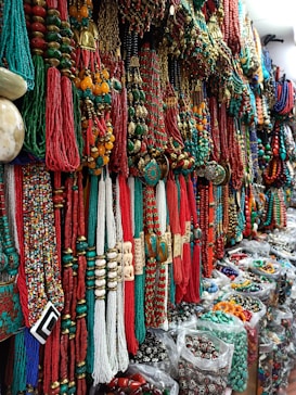 A vibrant display of colorful necklaces and beads, with shades of red, green, and gold dominating the intricate patterns. The necklaces are densely arranged, creating a rich tapestry of textures and materials. Located in a market setting, the variety and abundance of jewelry convey a sense of cultural richness.