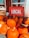 A display of bright orange pumpkins arranged outside a store, accompanied by a red sign with the word 'LOCAL' in white letters. Some bags are visible next to the pumpkins, along with a smaller sign indicating the price of firewood.