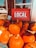 A display of bright orange pumpkins arranged outside a store, accompanied by a red sign with the word 'LOCAL' in white letters. Some bags are visible next to the pumpkins, along with a smaller sign indicating the price of firewood.