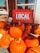 A display of bright orange pumpkins arranged outside a store, accompanied by a red sign with the word 'LOCAL' in white letters. Some bags are visible next to the pumpkins, along with a smaller sign indicating the price of firewood.