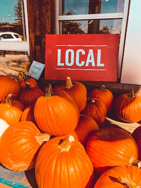 A display of bright orange pumpkins arranged outside a store, accompanied by a red sign with the word 'LOCAL' in white letters. Some bags are visible next to the pumpkins, along with a smaller sign indicating the price of firewood.