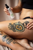 A person is holding a bottle of tattoo care product labeled 'Stories & Ink' above a tattoo on their leg. The tattoo features vibrant images with text that appears to say 'WORKERS UNITE' surrounded by colorful designs and characters.