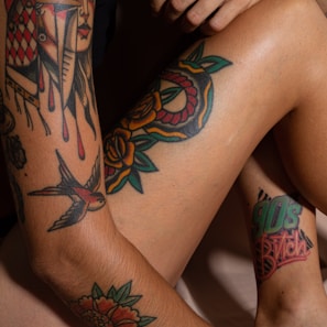 Various colorful tattoos are displayed on skin, including a geometric face design, flowers with bold outlines, and a bird in flight. The word '90s Bitch' is inked on the leg in a vibrant font.