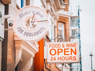 A street scene featuring two signs. The first sign is a neon clock shaped like a doughnut with the text 'Always a good time for doughnuts.' The second sign is rectangular with bold text advertising 'FOOD & WINE OPEN 24 HOURS' against an urban backdrop of buildings with brick and classical stone architecture.