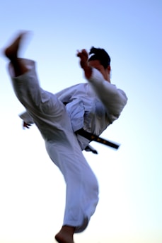 A person in a white martial arts gi executing a high kick against a clear blue sky. The figure is in mid-air, showcasing a dynamic and powerful pose, with movement captured in the focus.
