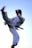 A person in a white martial arts gi executing a high kick against a clear blue sky. The figure is in mid-air, showcasing a dynamic and powerful pose, with movement captured in the focus.