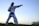 A person wearing a traditional martial arts uniform with a black belt stands in a stance, poised with one arm extended in a punch. The background features an open sky transitioning from light to deep blue, suggesting early evening or dawn. The ground is grassy, and some foliage is visible.