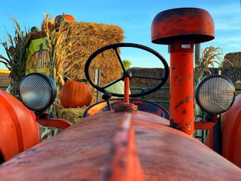A red tractor is prominently featured in the foreground, with a round steering wheel centered above the body. On either side of the tractor, large circular headlights are visible. In the background, a stack of hay bales is decorated with pumpkins and corn stalks. A sign for 'Kat Acres Farm' is partially visible, adding an autumnal farm setting.