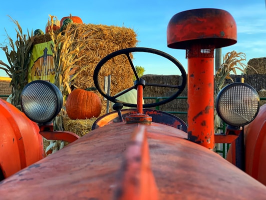 A red tractor is prominently featured in the foreground, with a round steering wheel centered above the body. On either side of the tractor, large circular headlights are visible. In the background, a stack of hay bales is decorated with pumpkins and corn stalks. A sign for 'Kat Acres Farm' is partially visible, adding an autumnal farm setting.