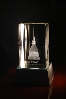 A clear glass block with an engraved image of a building featuring a tall spire, placed on a dark, reflective surface with a strong light illuminating from one side. The word 'TORINO' is etched below the image.