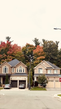 Two suburban houses with brick facades are located side by side. The homes have well-manicured lawns and driveways, each with a vehicle parked. Autumn trees with vivid red, orange, and green foliage are visible in the background, adding a seasonal touch.