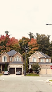 Two suburban houses with brick facades are located side by side. The homes have well-manicured lawns and driveways, each with a vehicle parked. Autumn trees with vivid red, orange, and green foliage are visible in the background, adding a seasonal touch.