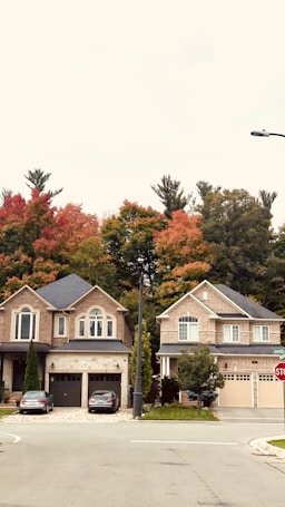 Two suburban houses with brick facades are located side by side. The homes have well-manicured lawns and driveways, each with a vehicle parked. Autumn trees with vivid red, orange, and green foliage are visible in the background, adding a seasonal touch.