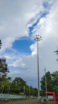 Tall pole with multiple lights stands under a partly cloudy sky, surrounded by green trees and a road running beside a painted wall.