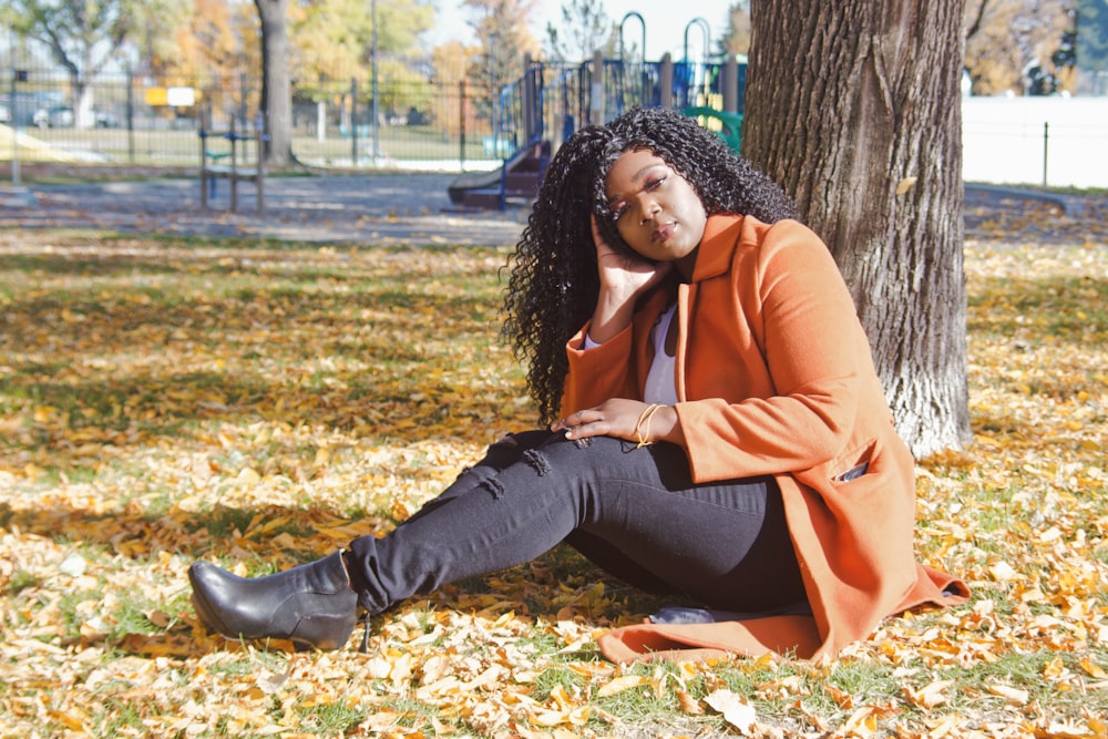 woman in brown leather jacket and blue denim jeans sitting on ground with dried leaves during