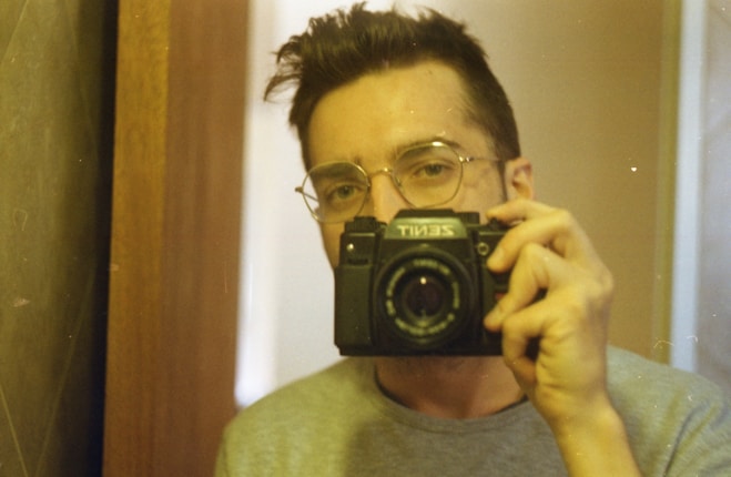 A person with short dark hair and glasses holds an old-fashioned Zenit camera up to their face, capturing a self-portrait in a mirror.