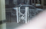 A chain and padlock secure metal handles on a glass door, with a reflection of a parked car and street scene visible in the background. The atmosphere appears quiet and subdued.