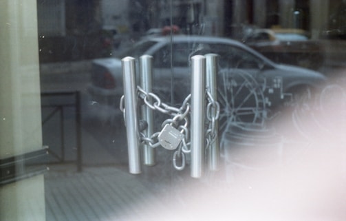 A chain and padlock secure metal handles on a glass door, with a reflection of a parked car and street scene visible in the background. The atmosphere appears quiet and subdued.