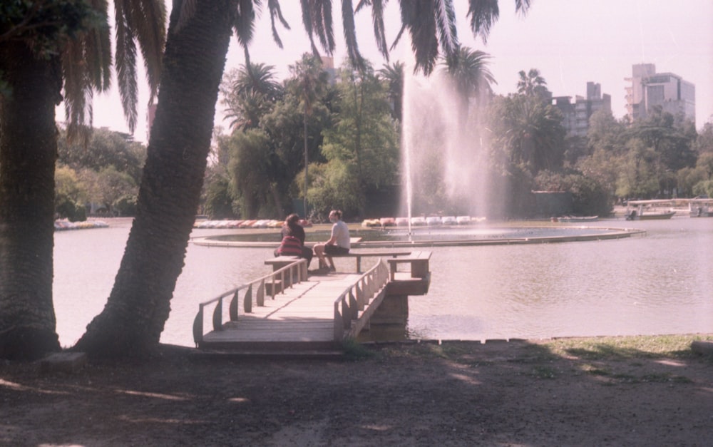 2 person sitting on brown wooden bench near water fountain during daytime