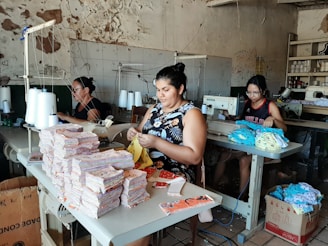A group of people work at sewing machines in a workshop with worn and peeling walls. The workspace is filled with stacks of fabric pieces, spools of thread, and sewing equipment. The individuals are focused on their tasks, handling different materials.